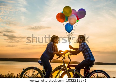 two people on romantic date looking each other. Young couple sitting at one bicycle.Girl holding colorful balloons on sunset cloudy sky background. man and woman. boy and girl on romantic date
