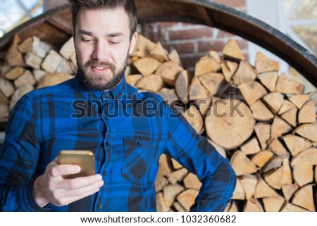 Smiling bearded hipster guy reading text message in social network via mobile phone standing against firewood design background with copy space. Cheerful businessman chatting on cell telephone