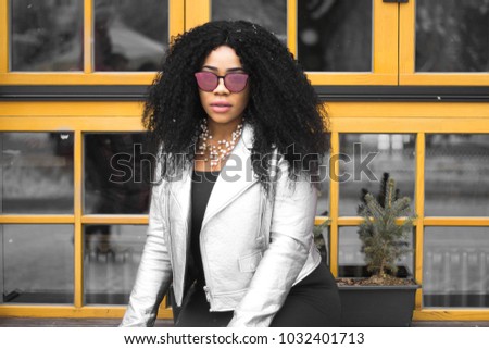 A young and stylish dark-skinned girl in a sunglasses with curly hair and silver jacket sitting in a spring city