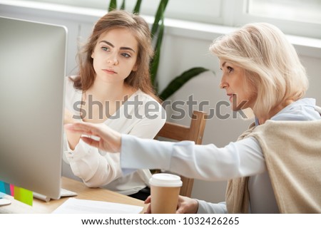 Senior and young female colleagues discuss online project look at pc screen, older mentor teaches young woman explains corporate software work, aged executive helps intern, teamwork on computer task