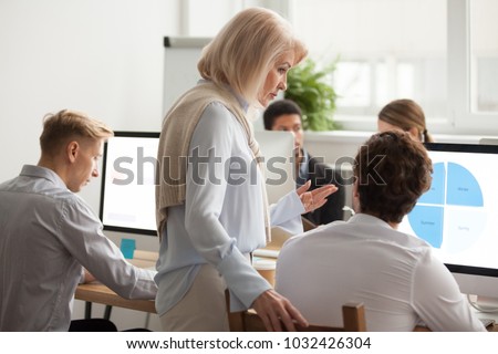 Senior female executive boss supervising computer work of young employee, serious aged woman mentor teaching intern explaining statistics data analysis, older leader helping colleague with problem Royalty-Free Stock Photo #1032426304