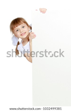Funny little blonde girl with pigtails and white bows on her head in a white T-shirt and shorts.She hid behind the white banner ad and peeks out because of him.