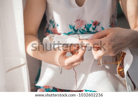 Young beautiful woman is engaged in embroidering a dagger on the embroidery frame in a colored dress at the window in the sunlight. A great picture for a hobby