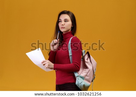 Photo of natural attractive journalist on an isolated orange background, wearing red sweater with throat, looking away. The correspondent is reporting and records the intriguing events in the notes.
