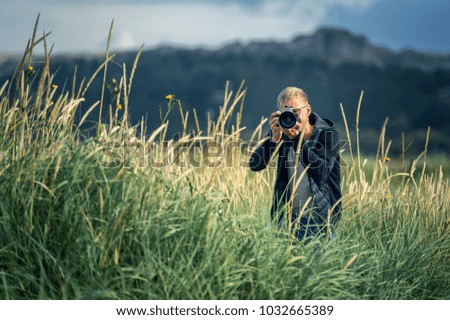 Portrait of young man in glasses standing among the high grass and taking a picture. Clouds on background. Norway.