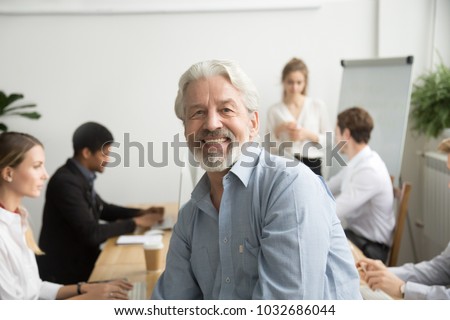 Smiling male senior team leader, aged teacher looking at camera with office people at background, happy old gray-haired company boss, experienced mentor or executive professional head shot portrait Royalty-Free Stock Photo #1032686044