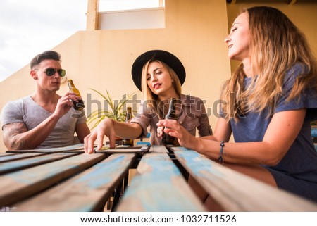 two young beautiful blonde woman and a man drinking beer on the terrace outdoor. Checking the messages on mobile smart phone on the wood table. friendship concept
