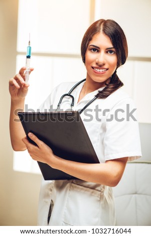Portrait of a beautiful young female nurse standing in her consulting room and holding injection. She is looking at camera with smile on her face.