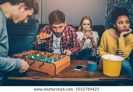 Two boys are playing board football game. They are very concentrated on it while girls are watching in the phone and feel boring. They don't like this game at all.