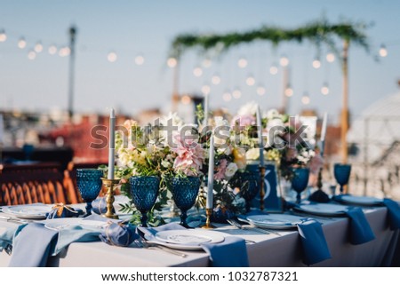 Long dinner tables covered with grey and blue cloth, served with porcelain and blue glasses and rich decorated with flowers stand on the roof of a house. Bokeh lights on baclground Royalty-Free Stock Photo #1032787321