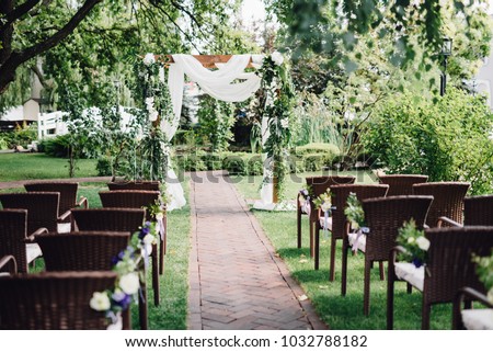 Wooden wedding arch  decorated by white cloth and flowers with greenery standing in the center of wedding ceremony. Brown chairs on side.