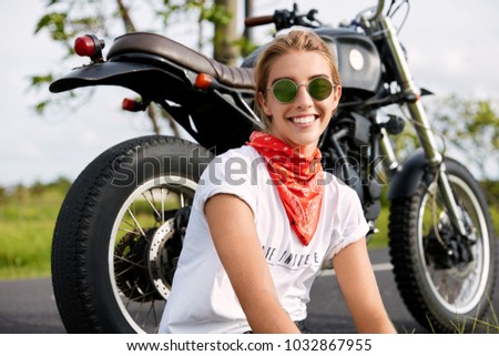 Portrait of cheerful female biker feels free and independent, stops on way with motorbike, enjoys tour journey in countryside, being in high spirit after active drive. People, lifestyle, hobby concept