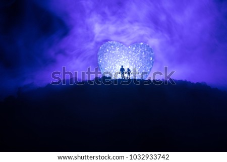 Silhouette of happy couple standing behind big shaped heart symbol on mountain at night. Big heart like moon glowing at foggy sky. Valentine`s day decor photo. Toned background