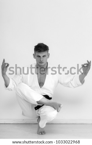 man, karate fighter or athlete, sitting in relaxing, yoga position with mudra hands, barefoot in kimono, suit, with black belt on white wall. Sport and meditation. Physical and mind training
