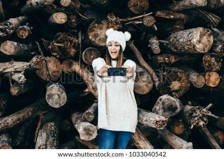 Young and pretty woman using her mobile phone outdoors, having fun taking herself pictures on a winter day with a background of wooden logs. Lifestyle.