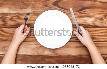 Hungry child waiting for meal. Child's hands holding fork and knife over table with empty dish. Top view. Royalty-Free Stock Photo #1033046173
