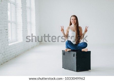Attractive girl with long brown hair sitting on metal black box and showing peace or victory on white studio background