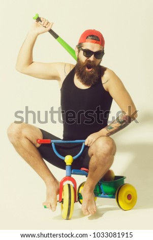 bearded man, long beard, brutal caucasian hipster with moustache in black vest, sunglasses, red cap holds green baseball bat with happy face sitting on colorfyl bicycle toy on grey background
