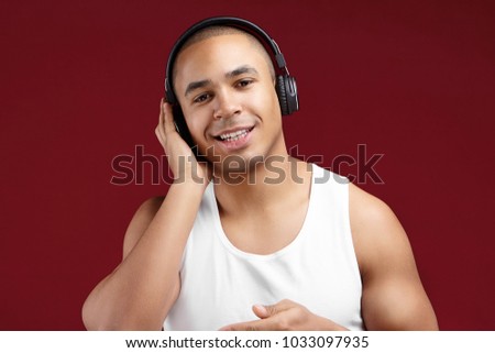 Cropped shot of handsome charismatic young dark skinned male DJ in white sleeveless t-shirt smiling joyfully, holding hand on his ear wearing wireless headphones while choosing tracks for party