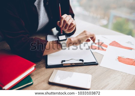 Cropped image of student checking time on display of modern wristwatch on hand while sitting at desktop with digital smartphone and design sketches for fashion collection.Smartwatch with blank screen