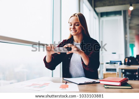 Cheerful blonde female designer making photos of fashion sketches on modern smartphone sitting at desktop in office.Positive young woman taking pictures on mobile phone working in stylish studio