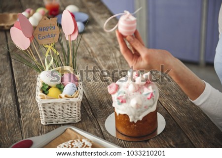Close up of female hand holding tinted egg. Focus on sweet easter cake and box with attributes on wooden table