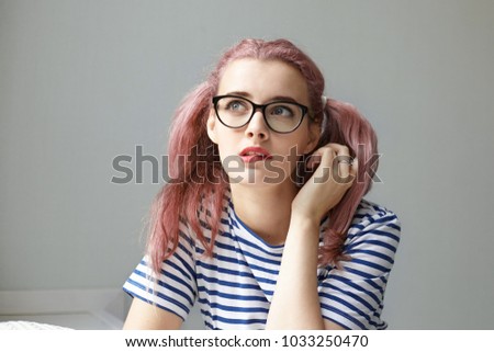 Portrait of beautiful Caucasian school girl with pinkish ponytails wearing stylish eyeglasses and sailor shirt doing homework and biting lips, having pensive expression, dreaming about something