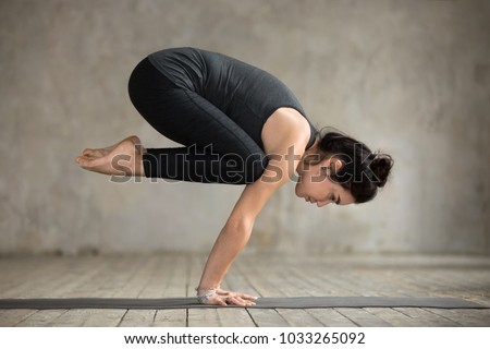 Young woman practicing yoga, doing Crane exercise, Bakasana pose, working out, wearing sportswear, black pants and top, indoor full length, gray wall in yoga studio Royalty-Free Stock Photo #1033265092