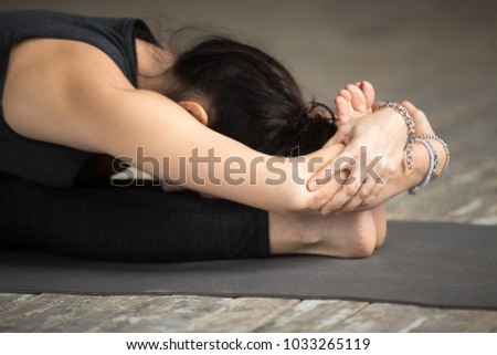 Young woman practicing yoga, doing paschimottanasana exercise, Seated forward bend pose, working out, wearing sportswear, black pants and top, indoor, gray wall in yoga studio, close up