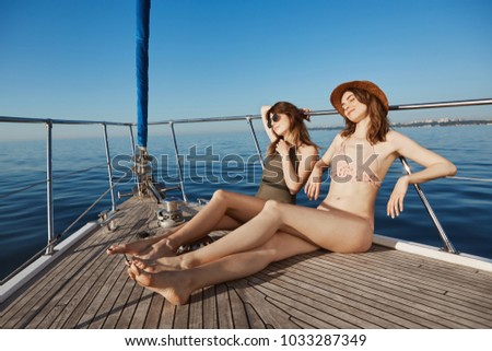 Two attractive adult woman on yacht, sailing in sea and sunbathing on bow of boat, feeling relaxed and pleased. Hot girls want get tan so they changed in bikinis. Summertime happiness
