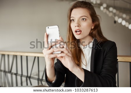 No one will believe me I saw it. Indoor shot of amazed and surprised woman sitting in cafe, holding smartphone while taking picture of celebrity who sits next to her, being shocked