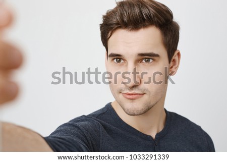 Studio shot of attractive cute man with stylish haircut and pleases expression taking selfie on smartphone, standing over gray background. Social media fan uploads another picture of himself