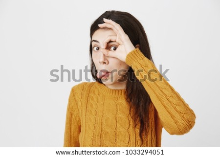 Funny cute caucasian girl sticking out tongue and showing okay sign over eye while looking through it as if it is telescope, standing over gray background. Kindergarten teacher plays with kids