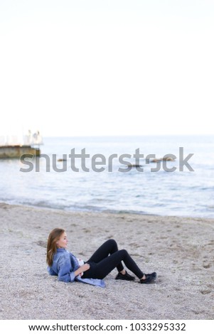 Happy girl looks at camera with beautiful and sweet smile, enjoying pleasant pastime and driving hands over warm sand with seashells, sitting on beach and looking toward endless blue sea on pleasant