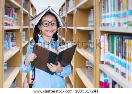 Picture of little girl looking at the camera with a book over her head while standing in the library