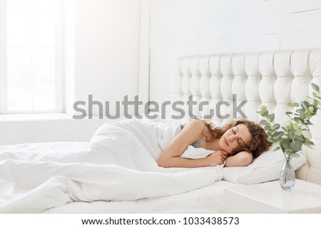 Horizontal shot of relaxed carefree female under white bedclothes in bed at bedroom, sees pleasant dreams, keeps eyes closed, enjoys good rest at home and calm atmosphere. People and rest concept