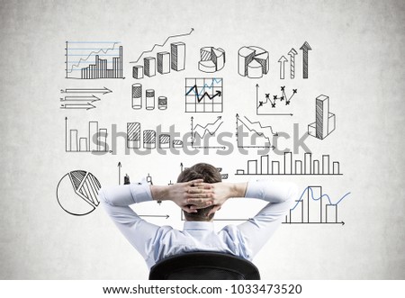 Businessman sitting in an armchair with hands behind his head looking at infographics on a concrete wall