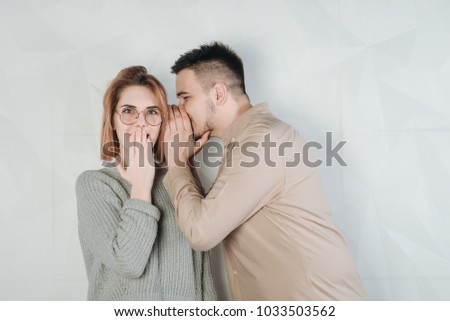 Man whispering a secret to a surprised young woman on white background. Bright picture of man and woman spreading gossip. Young guy whispers in ear the word girl and she laughs