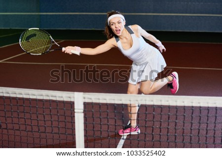 Full length portrait of forceful woman playing tennis in indoor court, swinging racket while hitting ball, copy space