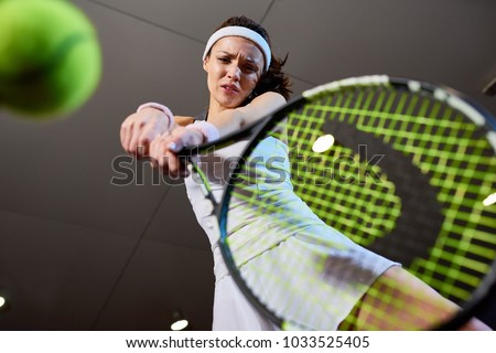 Low angle portrait of forceful woman playing tennis in indoor court, focus on tennis racket hitting ball, copy space