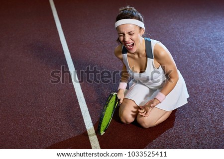 High angle portrait of female tennis player injured during practice screaming in pain sitting on court floor