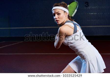 Waist up portrait of confident young woman playing tennis in dark indoor court, forcefully hitting ball with racket, copy space