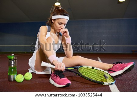 Full length portrait of sad young woman sitting on floor in indoor court resting after tennis practice, copy space