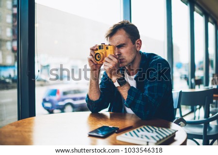 Pondering professional male photographer dressed in casual clothes trying to make good picture using quality equipment.Pensive handsome hipster guy taking photo using vintage yellow camera in cafe