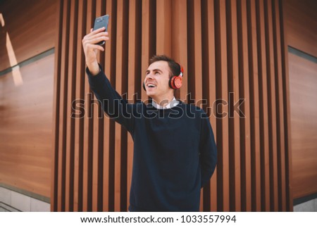 Good looking smiling male blogger making funny selfie on modern smartphone camera for sharing image with followers in network.Young cheerful man taking photo using cellphone while strolling outdoors