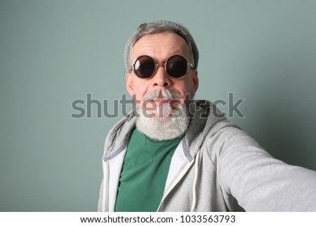 Happy senior man in hipster outfit taking selfie on grey background
