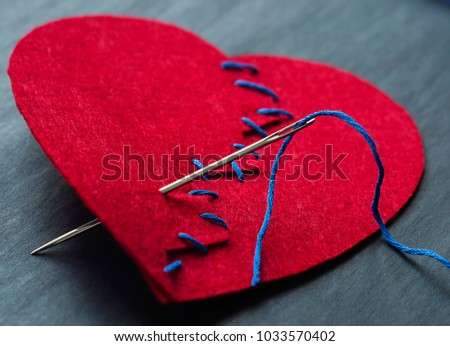 Symbolic image of broken heart. Textile red heart sewn with a needle and thread. Selective focus.