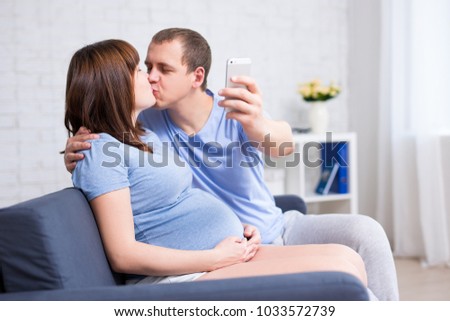 happy pregnant couple kissing and taking selfie photo with smart phone in modern living room