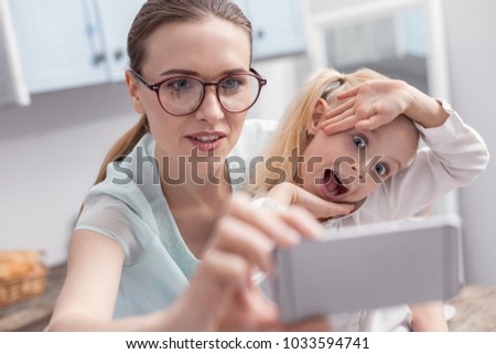 Free time. Charming young single mom putting on glasses while holding phone and daughter opening mouth