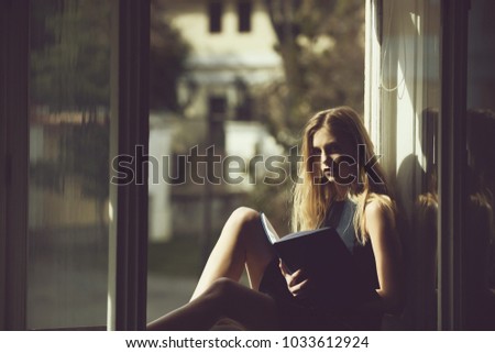 Pretty girl or young woman, student, teenager, with cute young face and blond, long hair in summer dress reading book at open window on sill on urban background. Leisure and relaxing